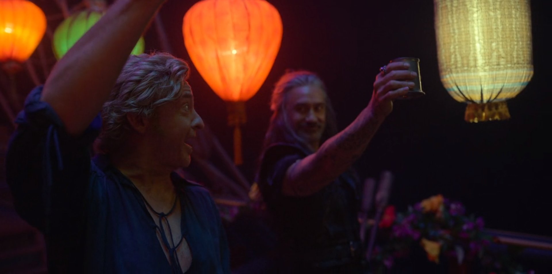 Photo still from the episode Calypso's Birthday. Stede and Ed are on the deck of The Revenge, with lanterns and decorations surrounding them. Stede holds his hand above his head and smiles broadly at Ed. Ed is also smiling, while holding his drink out in salutation.