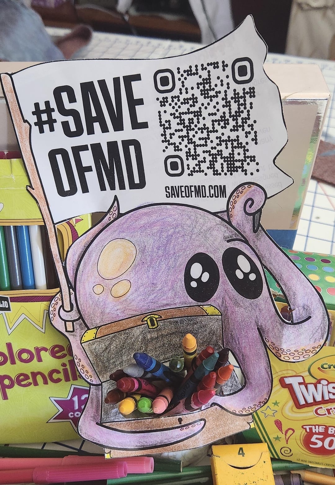 Flat Jeff is a cut out of an octopus, which has been colored with purple crayon. The octopus clutches a treasure chest filled with actual crayons and is surrounded by more packages of crayons and markers. One tentacle waves a flag with #SaveOFMD and a QR code to the petition on saveofmd.com.