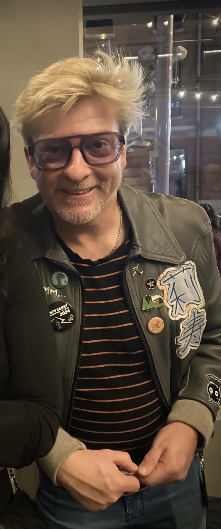 picture of Rhys Darby in leader jacket with patches and buttons, wearing purple glasses.