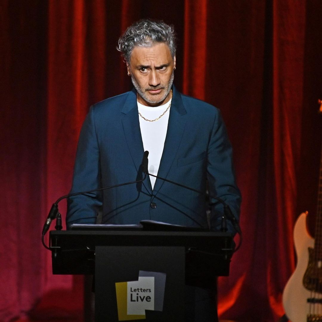 Picture of maori man, Taika Waititi reading at a podium in a blue suit with red curtains in the background