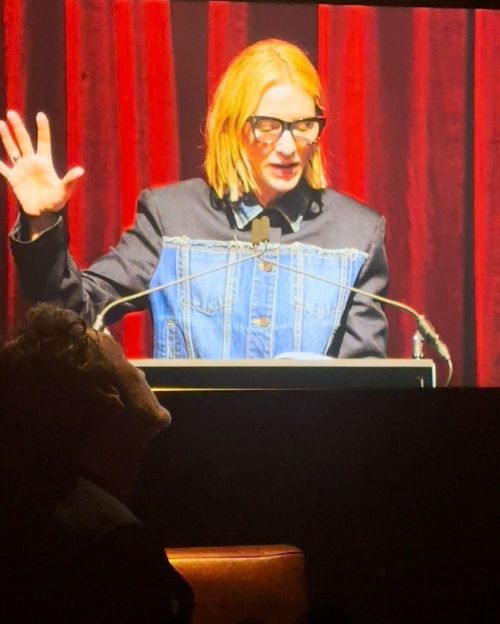 Picture of the back of taika waititi, reaching his arm up at Cat Blanchett on a projector.