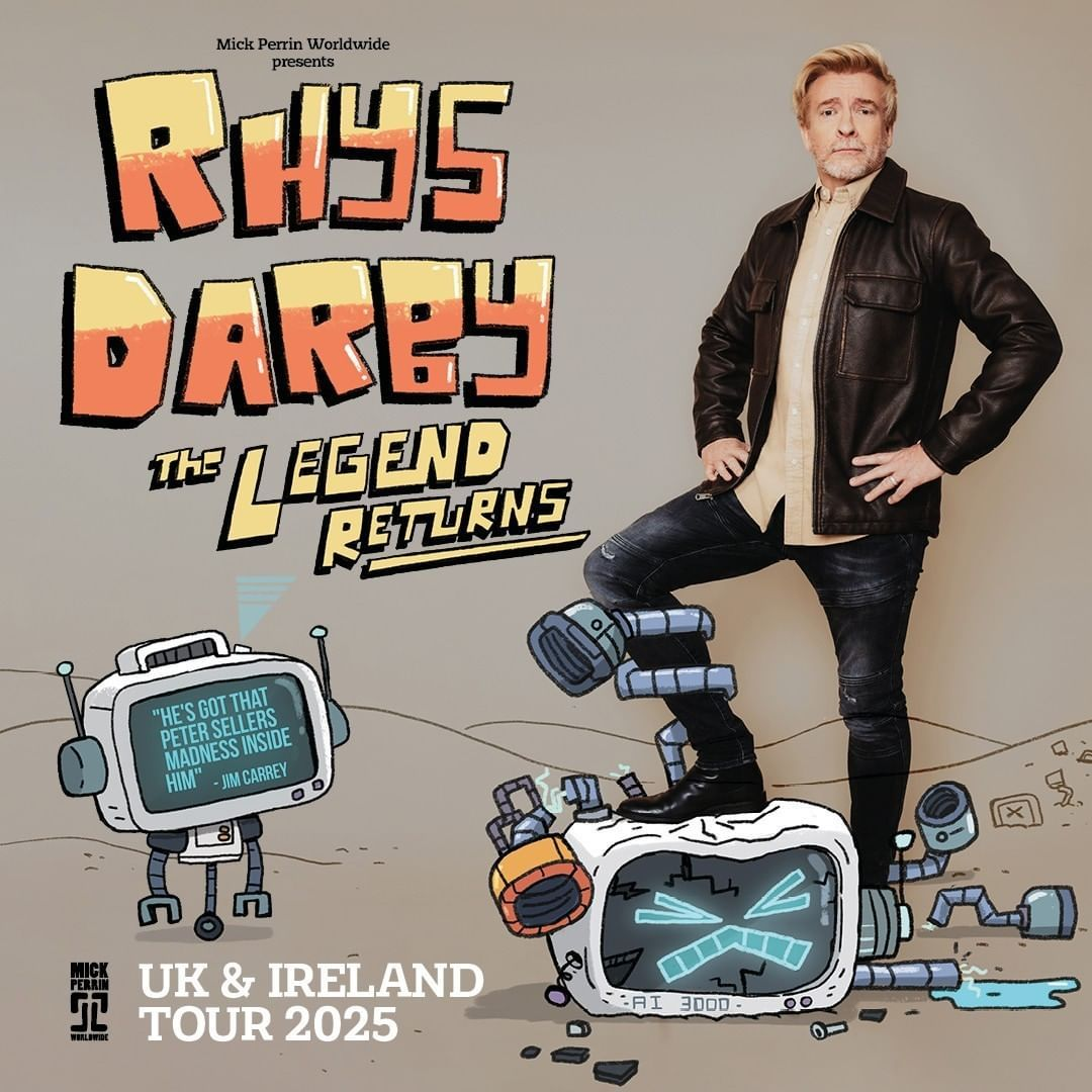 Rhys Darby stands in a white button down under shirt under a brown leather jacket and black jeans. He is standing with one leg up on a crushed cartoon robot.  In small letters, 