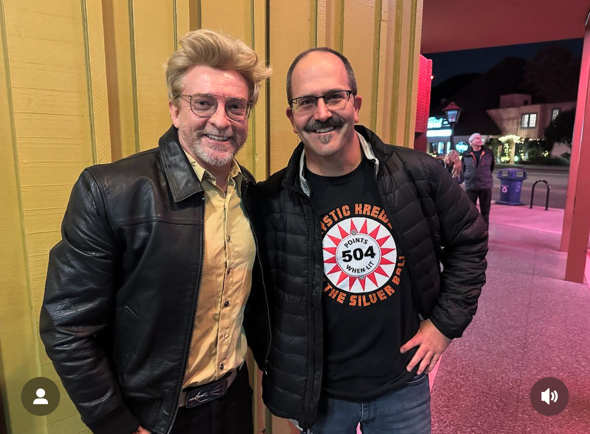 Rhys darby standing next to a fan with a yellow background. both smiling.