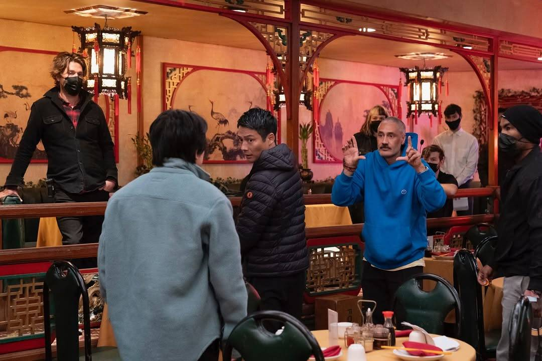 Photo of Taika Waititi on set with some of the Interior Chinatown cast and crew. The set is the inside of a Chinese restaurant. Taika is holding his hands up in a frame gesture while directing.