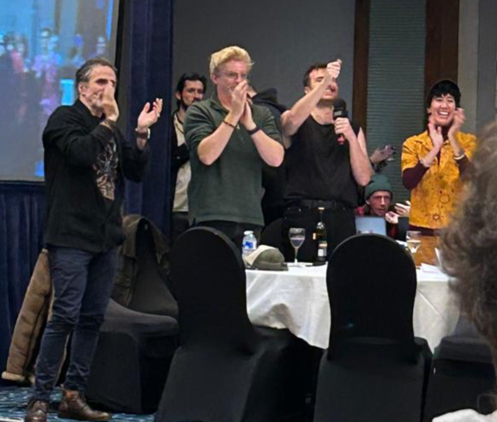 Picture of several cast members standing and clapping for the Cosplay Contest third place winner @unicorndeathrace. Pictured left to right is Con O'Neill, Rhys Darby, Nathan Foad, and Vico Ortiz with other people in the background.