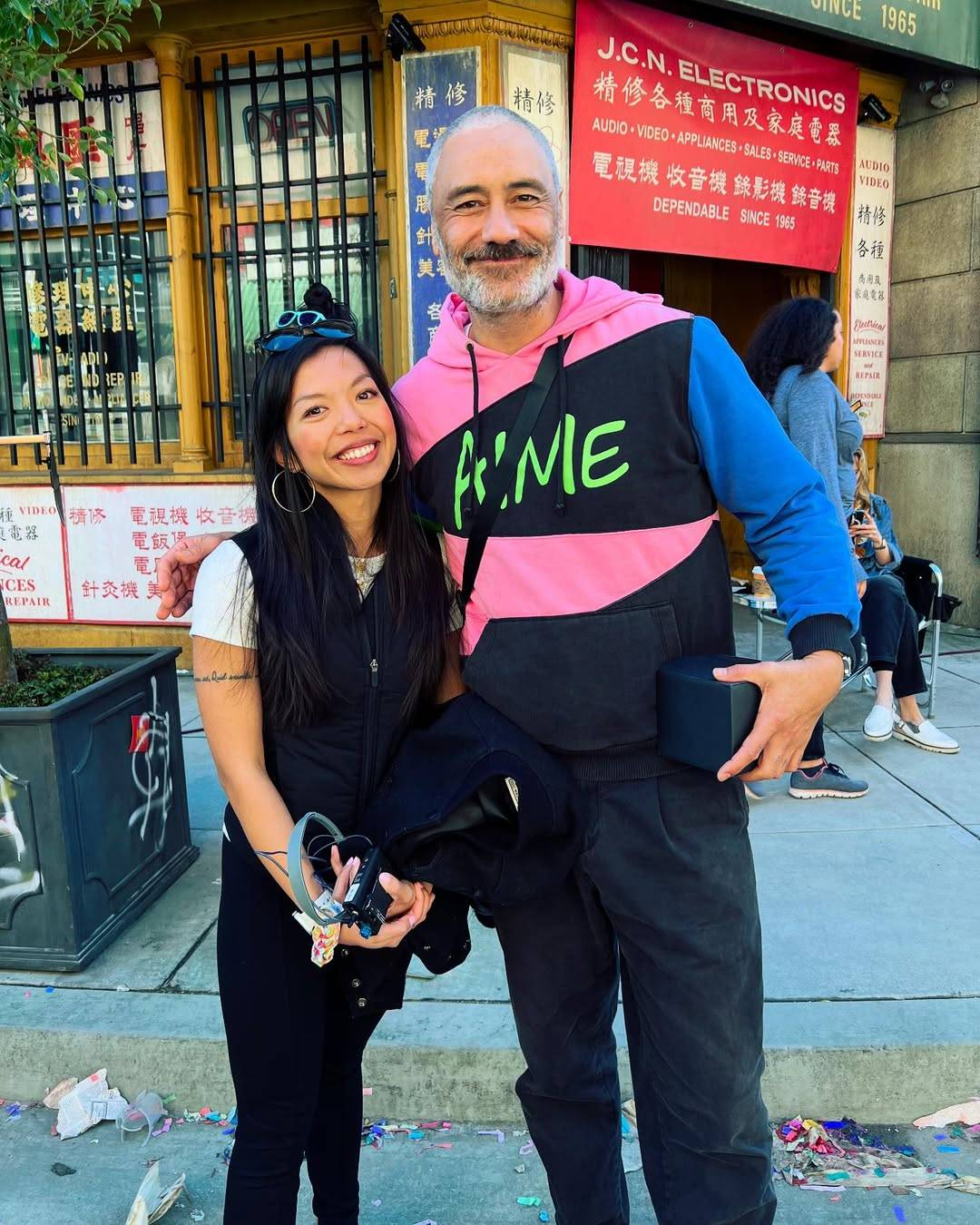 Photo from the set of Interior Chinatown. Taika and Judy Lei, the show’s dialect coach, are standing outside a Chinese electronics shop. Taika has one arm around Judy’s shoulders and both are smiling at the camera.