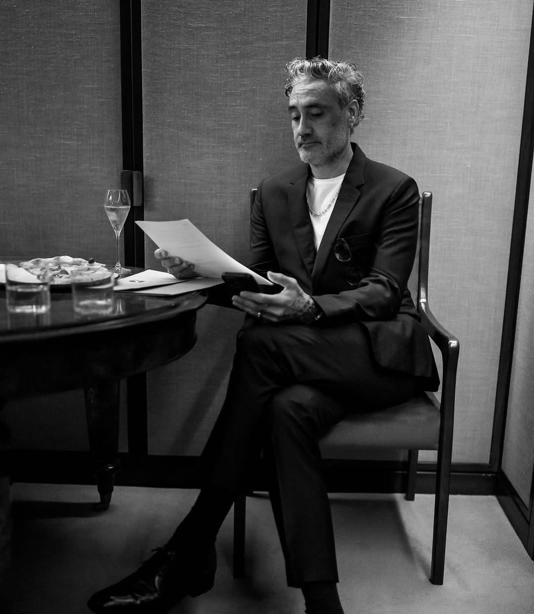 Black and white photo of Taika sitting at a small table and reading a letter while holding his phone in his other hand. He is dressed in a slim-fitting dark suit for the KokoFoundation event.