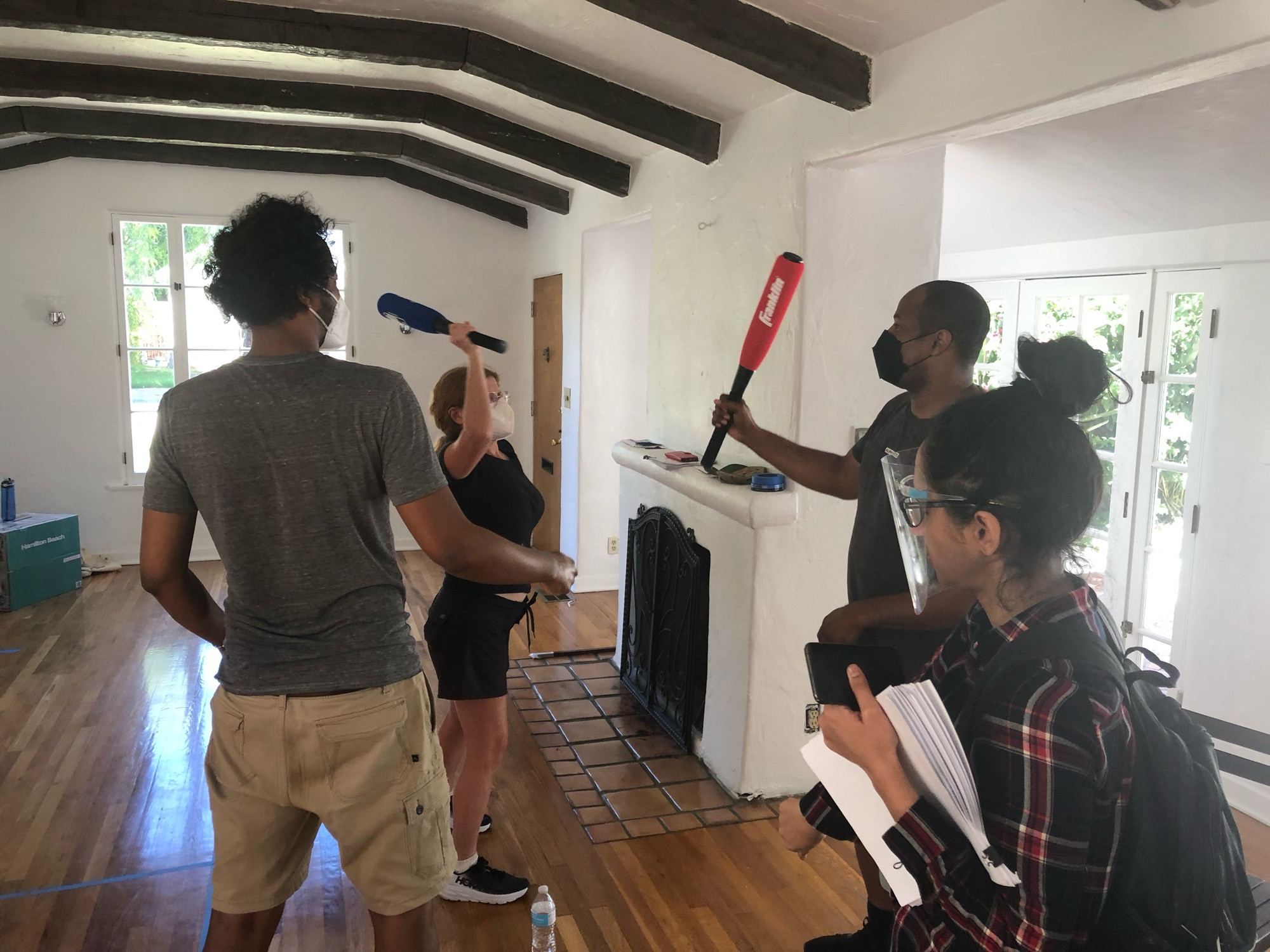 Photo of Samba on set with other cast/crew members. Samba is standing with his back to the camera, watching as a woman and a man appear to demonstrate or practice a fight, each of them holding a baseball bat.