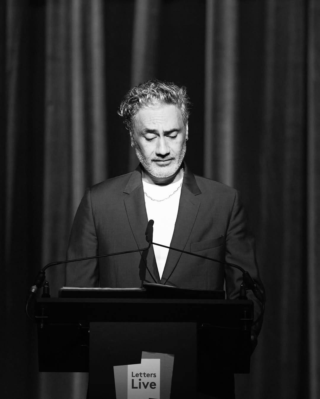 Black & white photo of Taika standing at a podium. He is looking down at his notes with a serious expression, his brow furrowed. He is wearing a dark suit jacket over a white t-shirt.