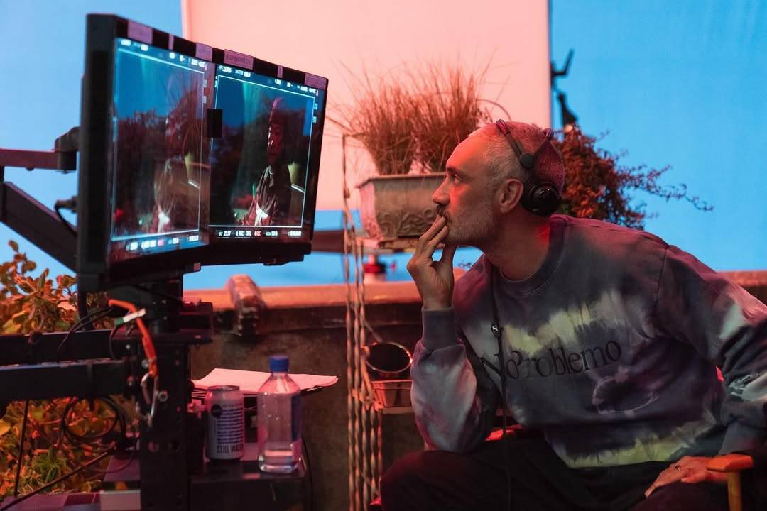 Photo of Taika directing an episode of Interior Chinatown. He is sitting in a chair and leaning forward with one hand on his chin while looking intently at a setup of two screens. In the background, we see a bluescreen and parts of the set, including a wall and some plants on a shelf.