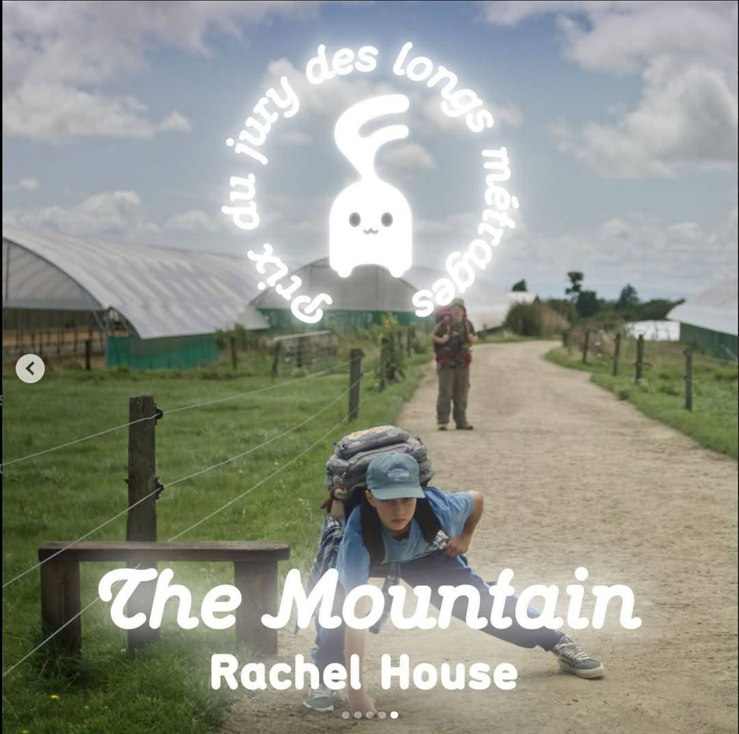 Image from the film, showing two children on what looks like a rural gravel road. Both are wearing big backpacks and the kid at the front is leaning down on one fist, with one leg bent, the other stretched out, while the other kid looks on from the background. “The Mountain – Rachel House” is written at the bottom of the picture, and the Festival Cinéma Jeune Public logo is at the top, a cute little mascot with the French text “Prix du jury des longs métrages” written in a circle around it.