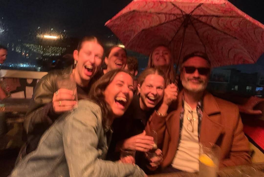 Taika Waititi and a group of five young women who are all laughing or smiling as they stand beside and behind him for the photo, drinks in hand. Taika himself is sitting down, holding an umbrella over himself and some of the group.