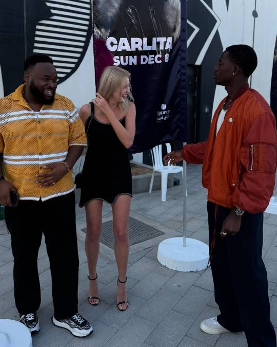 Photo of Samson Kayo with a woman and a man. The woman stands next to Samson, looking off to the left while fixing her hair. They are both smiling or laughing and Samson is looking at the other man, who stands facing  Samson and the woman.