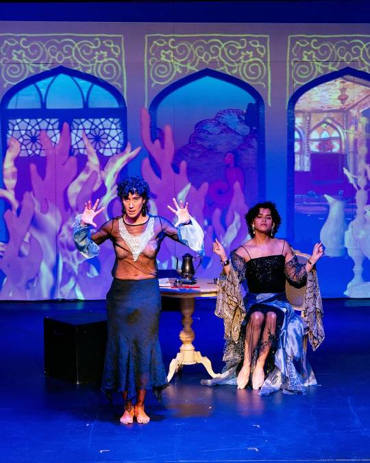 Vico and another cast member on stage. They are lit in blue, the backdrop showing flames licking up three large, ornately decorated windows or doorways. Vico stands in front, looking intensely at the audience with their arms raised, hands spread at about shoulder level. They are wearing a long skirt and a mesh top. The other cast member sits at a small round table slightly further back and to the right. They have about shoulder-length curly hair and are wearing long dangly earrings and a many-layered, fancy-lookimg dress with thin shoulder straps.