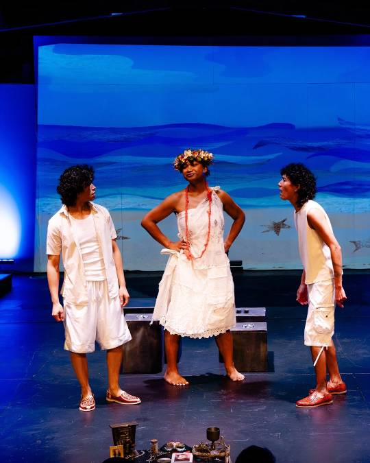 Vico and two other cast members, dressed in white, stand on stage in front of a painted backdrop showing a beach and the ocean. Vico is on the right and appears to be shouting at the person standing on the left, while the third cast member stands in the middle with their hands on their hips and a pleased or smug expression on their face.