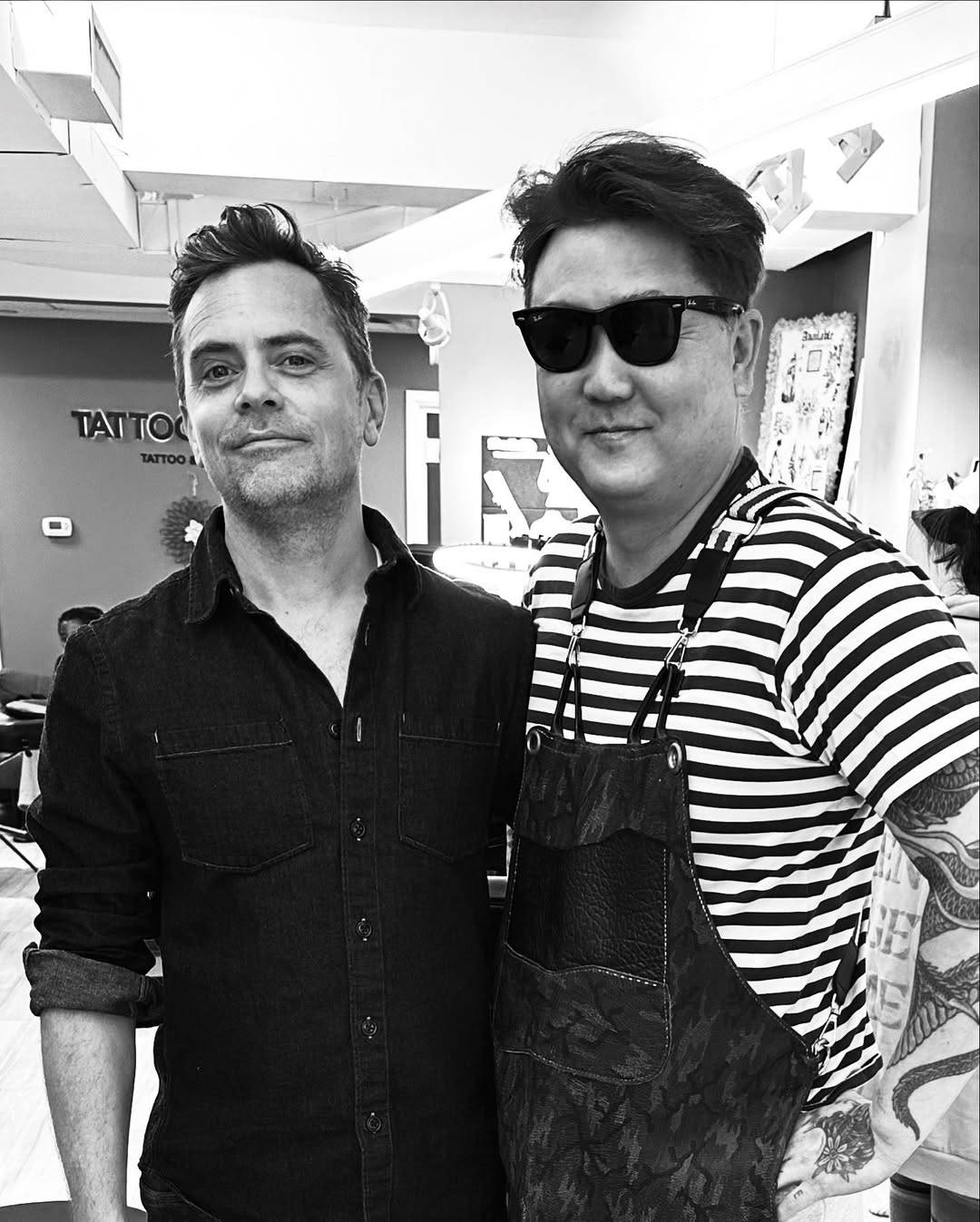 Black & white photo of David with his tattoo artist Chang. They are standing inside the tattoo studio with their arms on each other's back and smiling at the camera.