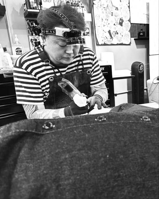 Black & white photo of Chang tattooing the inside of David's right arm. Chang is looking down in in concentration, wearing a headlamp, an apron and black rubber gloves.