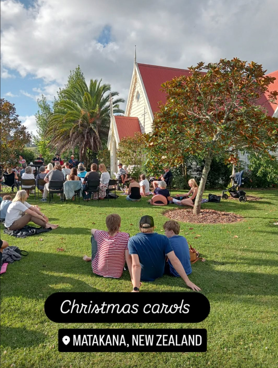 Rhys and his sons, seen from behind, sit on the grass and watch a band play at a small gathering outside a church. Small groups of people sit on jackets or blankets nearby, while others sit in plastic chairs closer to the church building. There are clouds in the sky but the pictured area is bathed in sun. Text at the bottom reads 