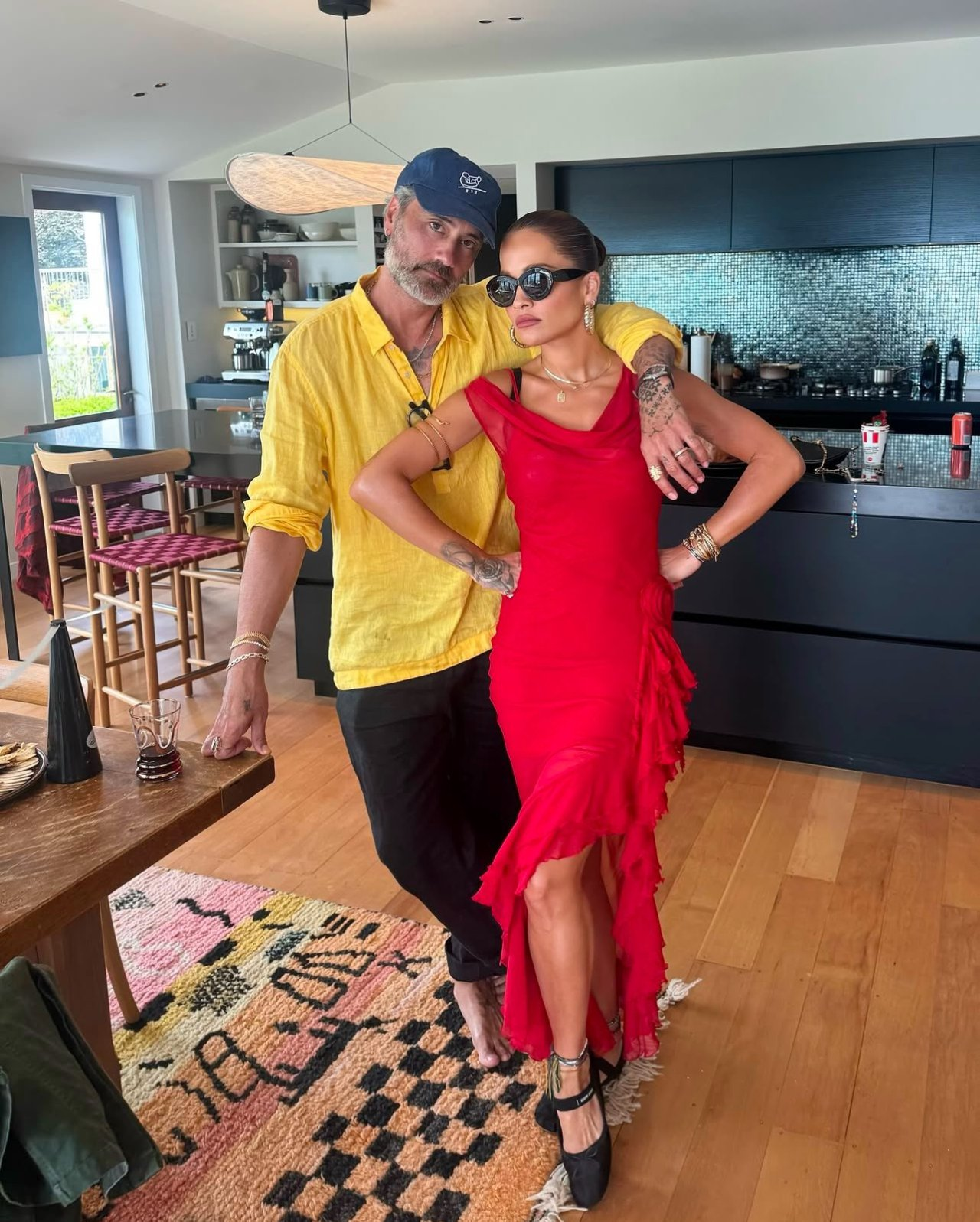 Taika Waititi and Rita Ora stand in a kitchen/dining room. Taika is leaning on a table with one hand, his other arm draped around Rita's shoulders. He is barefoot and wearing a long-sleeved but light yellow shirt with his glasses tucked into its collar, black trousers, and a blue baseball hat. Rita is wearing sunglasses, a red dress with a slit and frilly hem, and black ballerina-style shoes.