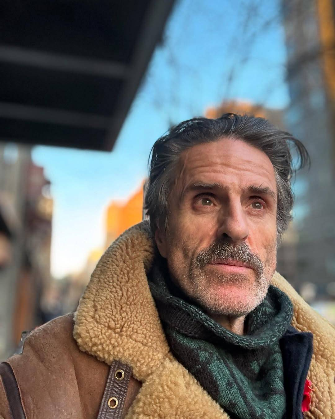 The accompanying photo is a selfie of Con, smiling slightly and looking at something just next to the camera. He is wearing a brown leather jacket with a fluffy collar, and a knitted scarf with a dark green and black pattern. Behind him, city buildings and a clear blue sky are visible but out of focus.