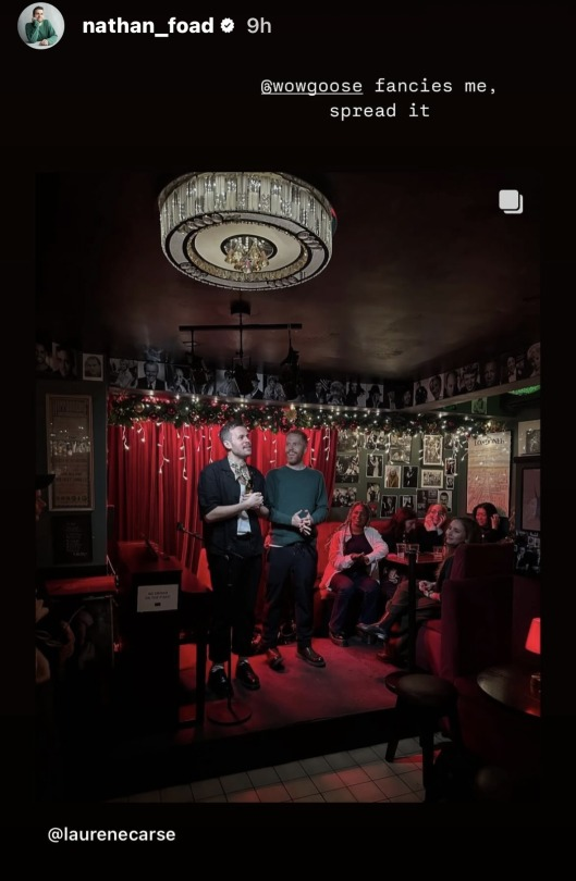 Insta story where Nathan shared a post from @ LaurenECarse, above which he writes: '@ WowGoose fancies me, spread it.' The post he shared is a photo of himself and another man standing on a tiny stage in a pub or bar. Nathan speaks as the other man watches, smiling broadly. Behind them is a red curtain, directly to their left is a piano, and to their right is a table, at which a small group of people sit and watch them. The walls around the stage are decorated with photos of many different people, and Christmas decorations hang from the ceiling.