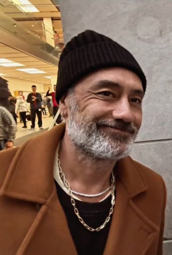 Photo of Taika wearing a black beanie and tan coat, giving a closed-mouth smile at the camera.