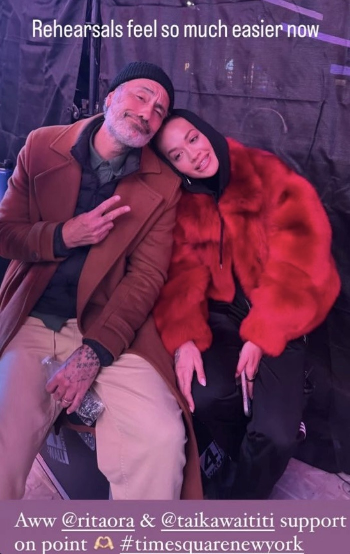 Taika and Rita sit on an equipment case ahead of the Times Square show. Rita is resting her head on Taika's shoulder and Taika's head is resting on hers. Rita is looking away from the camera and appears to be speaking, her mouth slightly open, while Taika smiles dopily at the camera with one hand raised to make a V shape with his fingers. Text at the top reads: 'Rehearsals feel so much easier now.' And at the bottom: 'Aww @ RitaOra & @ TaikaWaititi support on point (heart hands emoji, hashtag TimeSquareNewYork'.