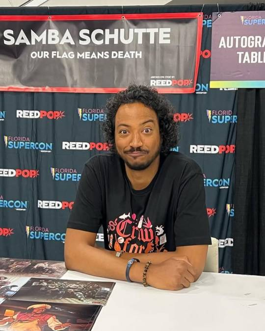 Photo of Samba sitting at his autograph table at Florida Supercon. He is wearing a 'Crew for Life' t-shirt and making a silly face at the camera.