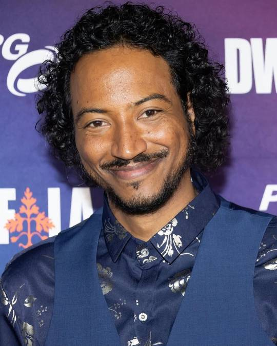 Close-up shot of Samba at a film premiere or event. He is wearing a dark blue waistcoat over a satiny-looking dark blue shirt with silver floral details, and he is looking at the camera with a closed-mouth but broad smile.