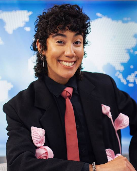 Portrait photo of Vico at a newsdesk in a black suit with pink roses pinned to it and a red tie, grinning at the camera.