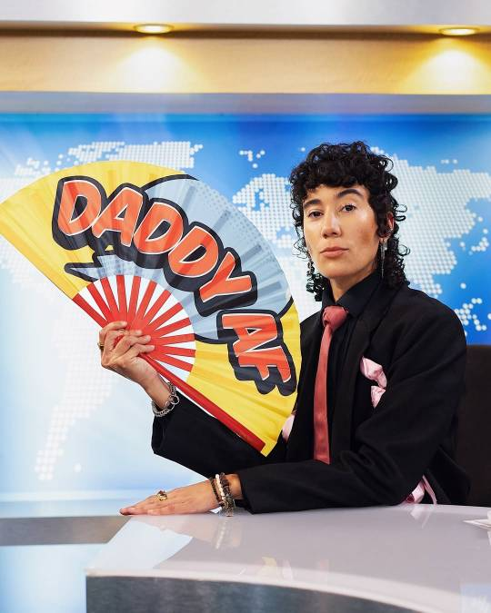 Vico at the newsdesk in their black suit and red tie, looking confidently at the camera while holding a fan. On the fan, 