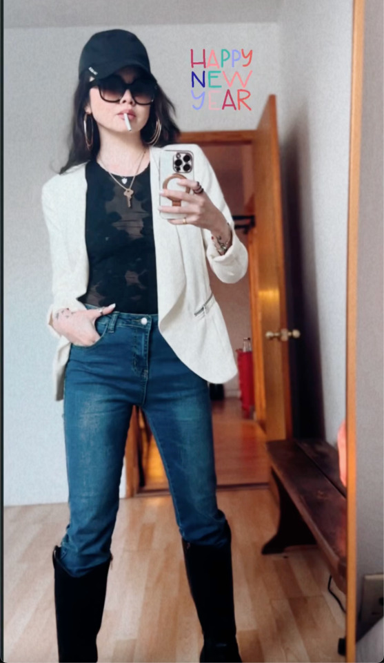 Full-body mirror selfie of Ruibo with an unlit cigarette between her lips, wearing a baseball hat, big sunglasses, a white blazer over a black top, blue jeans and black knee-high boots. She is holding up her phone with one hand, the other hand tucked casually into her jeans pocket. 'Happy new year' is written in colourful letters on the photo.