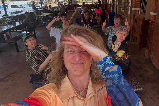 Photo of some of the Deadloch cast and crew outside a cafe or restaurant, taken by Kate Box, who is standing in the foreground and squinting at the camera with one hand shielding her eyes from the sun. The rest, including Madeleine, are in the background, giving thumbs up or otherwise gesturing at the camera.