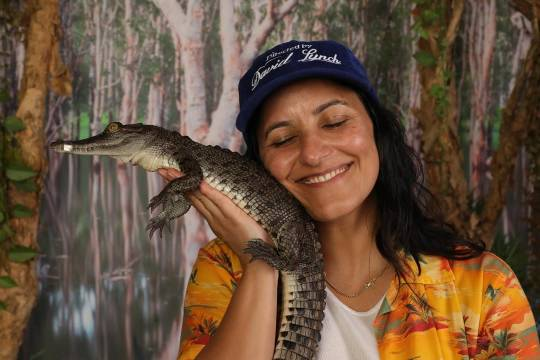 Close-up of Madeleine standing in front of a forest background, holding a baby crocodile or alligator up to her cheek. Her eyes are closed and she is smiling contentedly. 