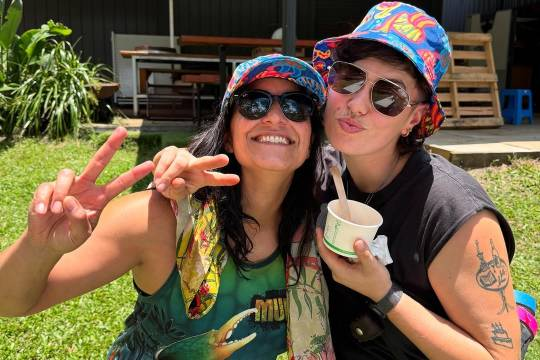 Madeleine Sami and a friend sit on the grass on a sunny day. They are both wearing sunglasses and similar but not identical blue hats with a colourful pattern. Madeleine is grinning at the camera and holding her hand up in a peace sign. Their friend is pouting at the camera with one arm draped over Madeleine's shoulder, also making a peace sign, while holding an ice cream cup in their other hand.