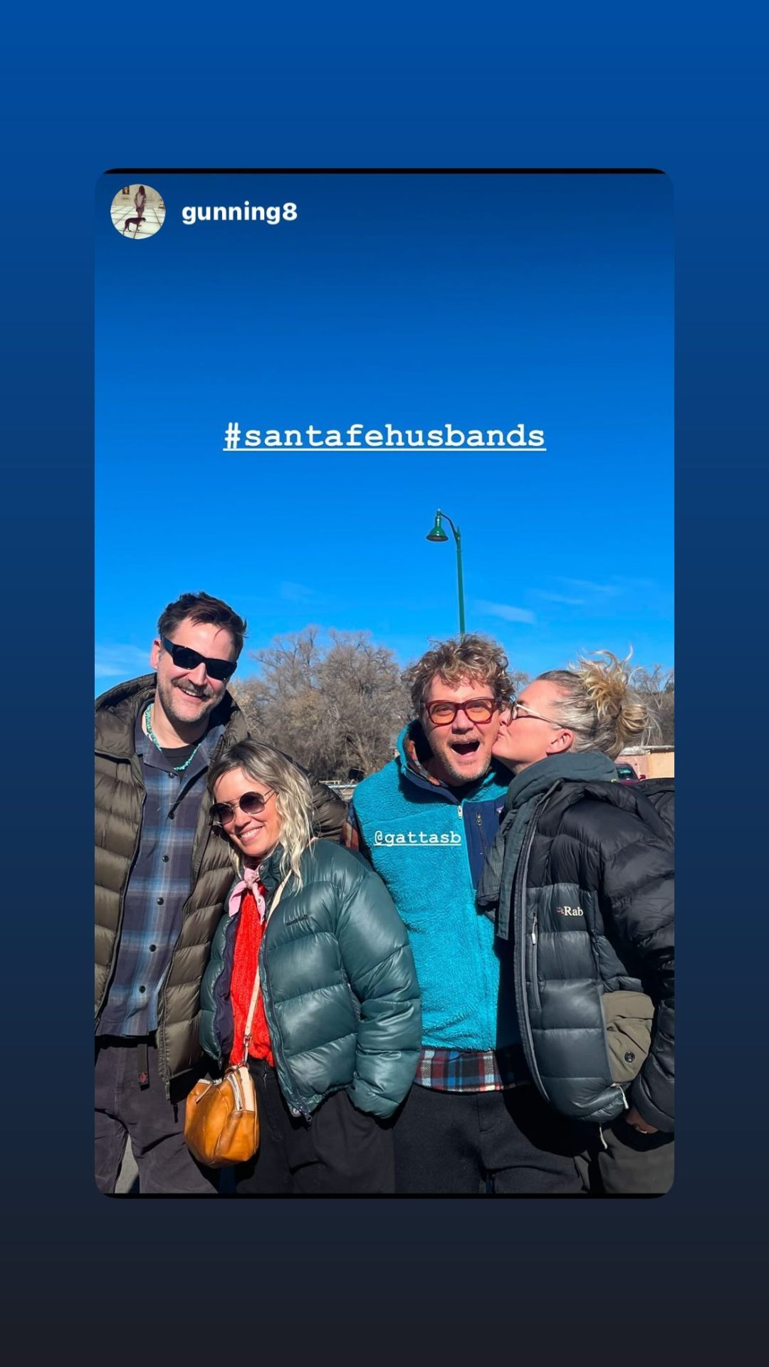 Insta story where Brian shared a post from @ gunning8: a photo of Connor and Brian with two women. They are standing outside on a sunny day with a clear blue sky, trees visible behind them. One woman is in a side-hug with Connor, while the other is kissing Brian on the cheek. ' # SantaFeHusbands' is written at the top of the photo.