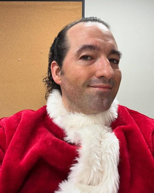 Selfie of Christopher, shot from the chest up. He is wearing the Santa suit already, but his face is bare and his hair slicked back. His head is turned slightly to the side but he is looking at the camera and smiling.