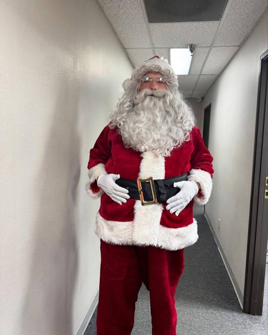 Christopher is now standing in a corridor and we can see the whole outfit from about shin height. He is resting his white-gloved hands on the wide black belt closed around his big Santa belly.