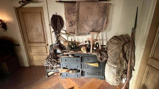 Photo of part of the set: a black dresser with a stool, maps, bottles, a skull, ropes and many other items strewn across it. Large map scrolls hang on the wall above it, and a big rifle leans against the wall next to it.