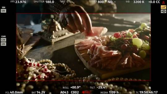 A camera screen shows the charcuterie board on Bartholomew's desk, with a pile of pearl necklaces and other jewelry next to it. Bartholomew's hand can be seen picking up some meat from the board.