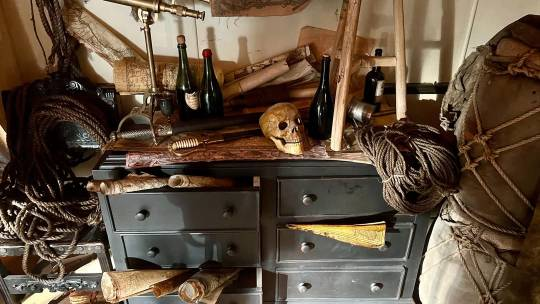 Close-up of the dresser, its drawers overflowing with paper scrolls. On top of the dresser, a telescope and some swords can be seen in addition to the items visible in the previous images. More ropes are piled onto a chair standing next to it.