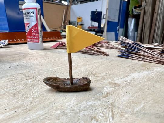 One of the tiny ships with a yellow triangular flag/sail sitting on a table in the workshop. Many more toothpick flags can be seen in the background, sorted by colour.