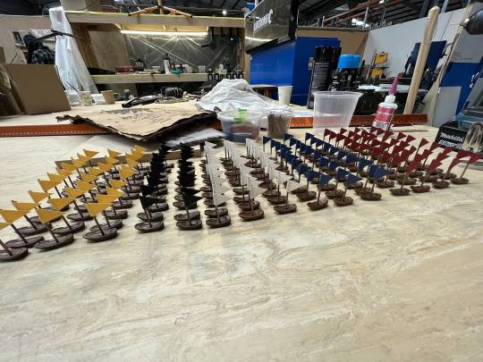 Rows of tiny boats lined up in the workshop, with yellow, black, white, blue and red flags.