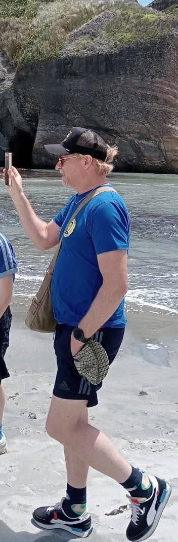 Photo of Rhys walking on a beach while holding up his phone. He is wearing a blue t-shirt, dark blue shorts and a black baseball hat. The hand not holding his phone is in his shorts pocket, and  there is another hat sticking out of said pocket.