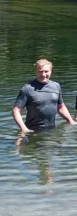 Photo of Rhys standing in the water, which is about hip height. He is looking at the camera and smiling. His t-shirt is soaked and plastered to his body.