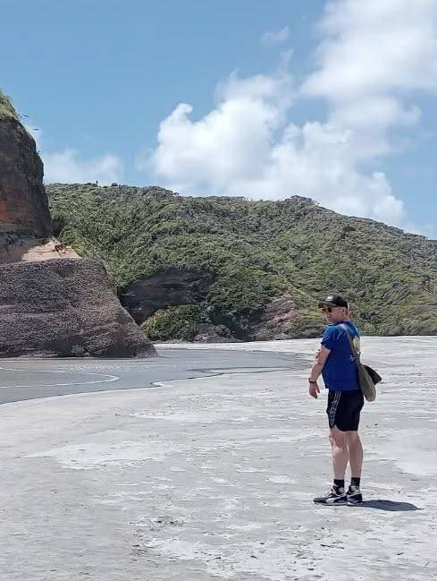 A wide shot of Rhys standing on a beach, facing a green hill and a large rock or outcropping. The photo is shot from behind him, and he is looking back over his shoulder and smiling. 