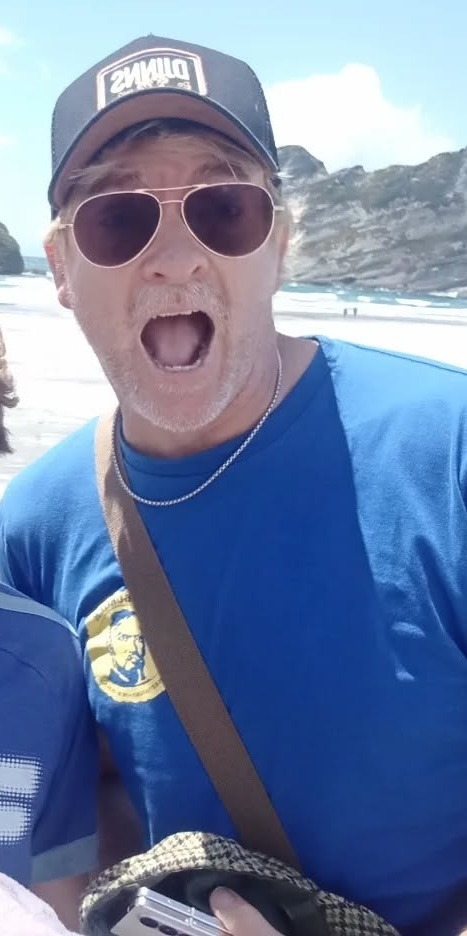 Another photo of Rhys at the beach. This one is up closer than the others, and he is making a funny face at the camera, with his mouth open and his eyes wide. He is wearing a pair of aviator sunglasses, a black baseball hat, and a blue t-shirt.