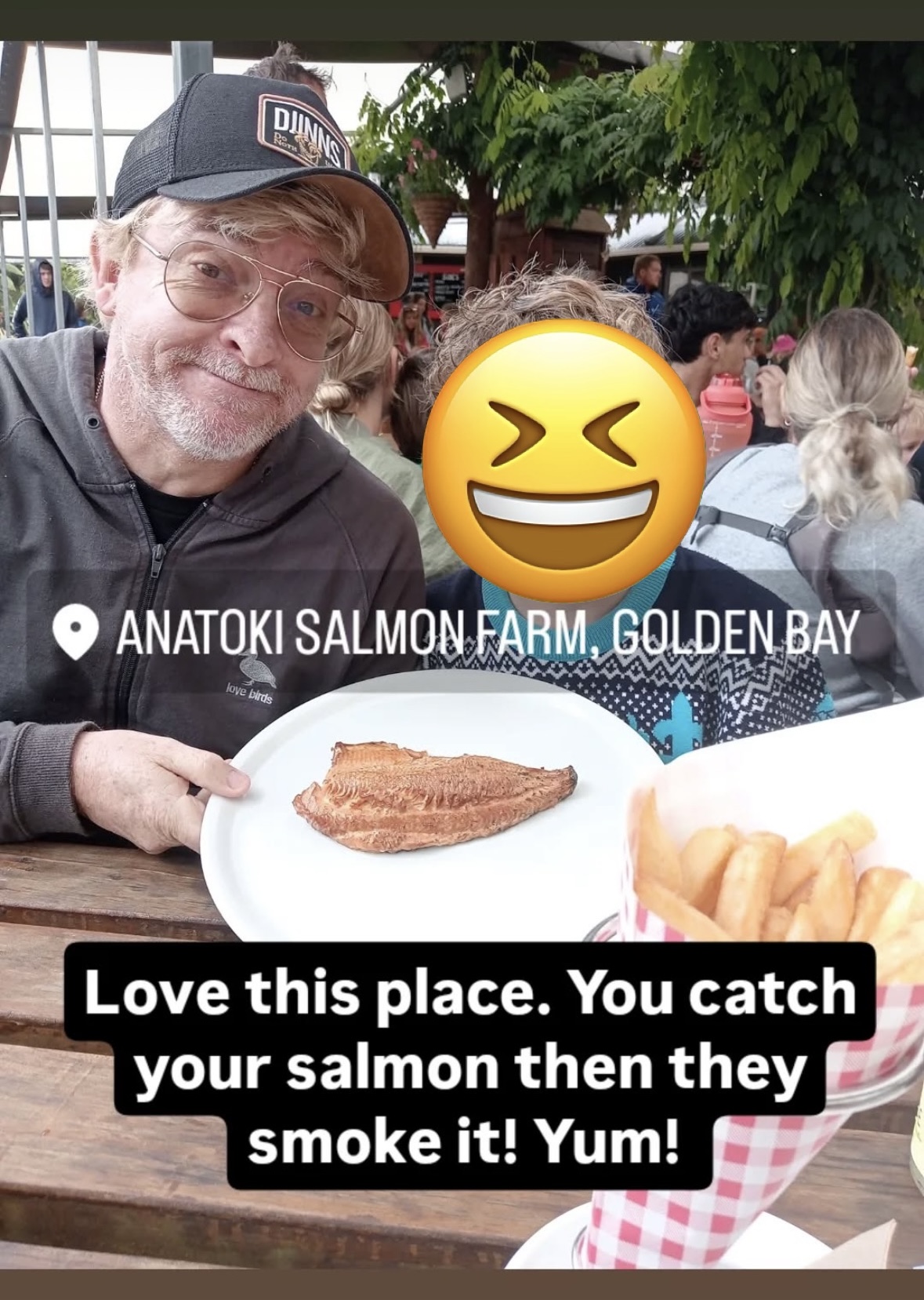 Rhys and one of his sons sit at a wooden table and show off their fish and chips. Rhys is smiling at the camera. His son's face is hidden by a laughing emoji. At the center of the photo, there is a location tag reading 'Anatoki Salmon Farm, Golden Bay' and at the bottom, text reads: 'Love this place. You catch your salmon then they smoke it! Yum!'