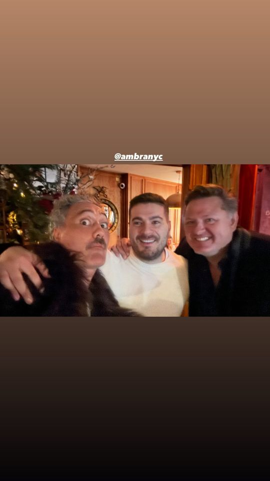 Instagram story with a photo of Taika and two other men at a restaurant. Taika is wearing a brown fur coat or collar and is staring at the camera with wide eyes, head turned slightly to the side and chin raised, while both of his friends are smiling. Above the photo, Taika has tagged @ AmbraNYC.