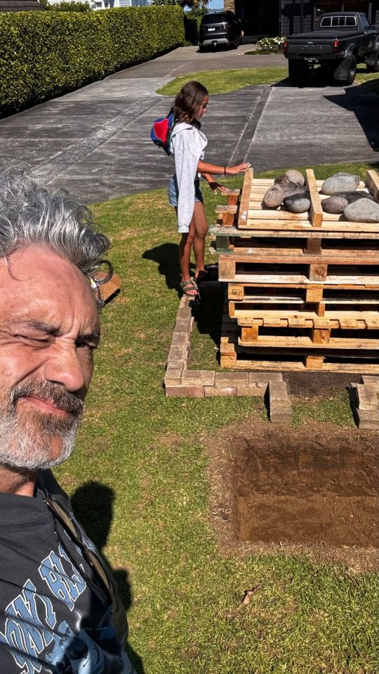 Selfie taken by Taika. He can be seen squinting at the camera in the bottom left corner, but the focus of the image is a stack of pallets with rocks on top. Bricks are laid out in a square around the pallets, and on the side facing the camera, they form a short channel to the hangi pit. Taika's daughter balances on the brick border, looking down at the rocks with her hands placed on the edges of the top two pallets.